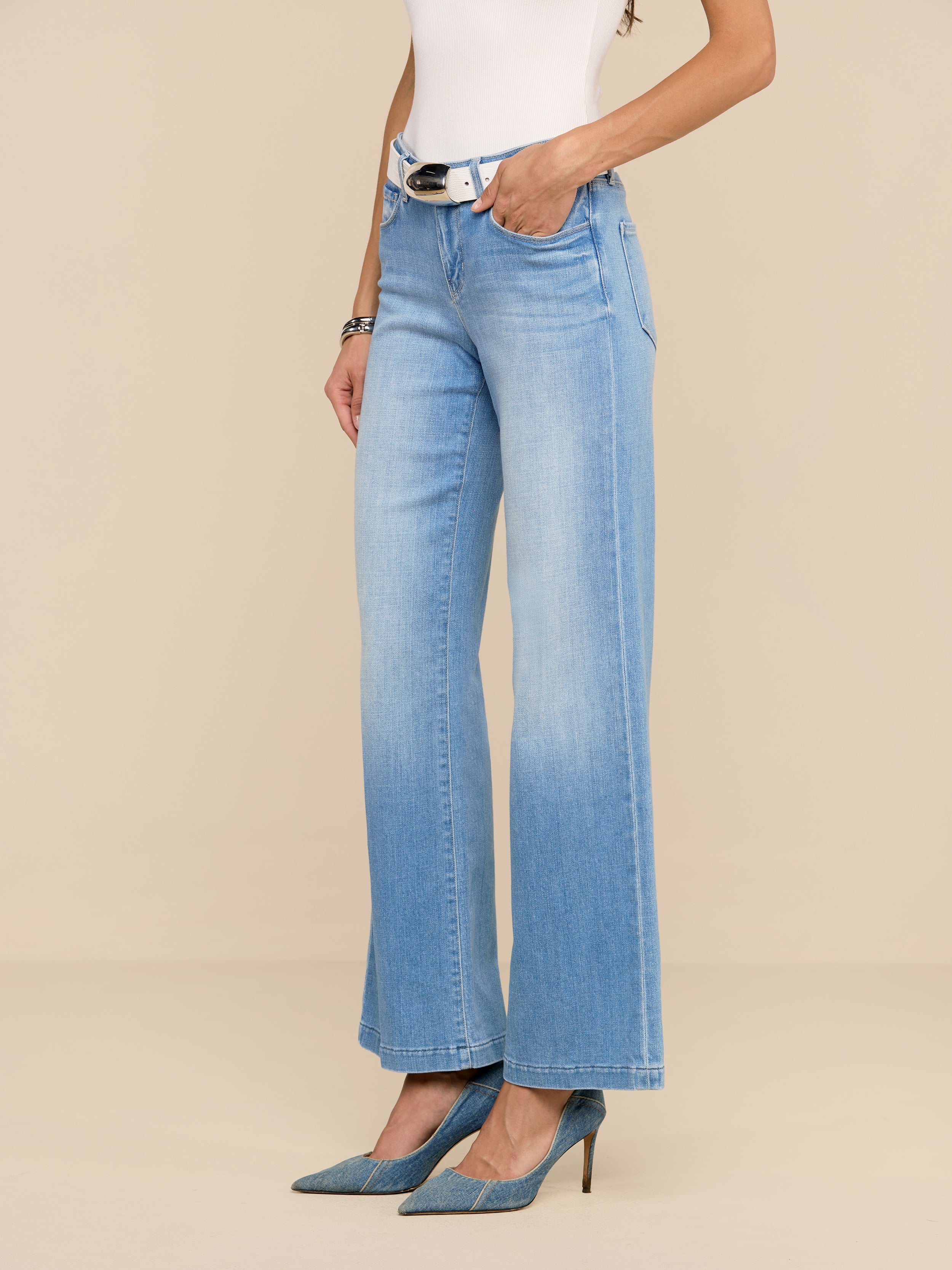 Une personne porte un jean Joanne à taille basse et à jambe large en délavage indigo clair, associé à une ceinture blanche, un haut sans manches blanc et des talons bleus à bouts pointus, sur un fond beige uni.