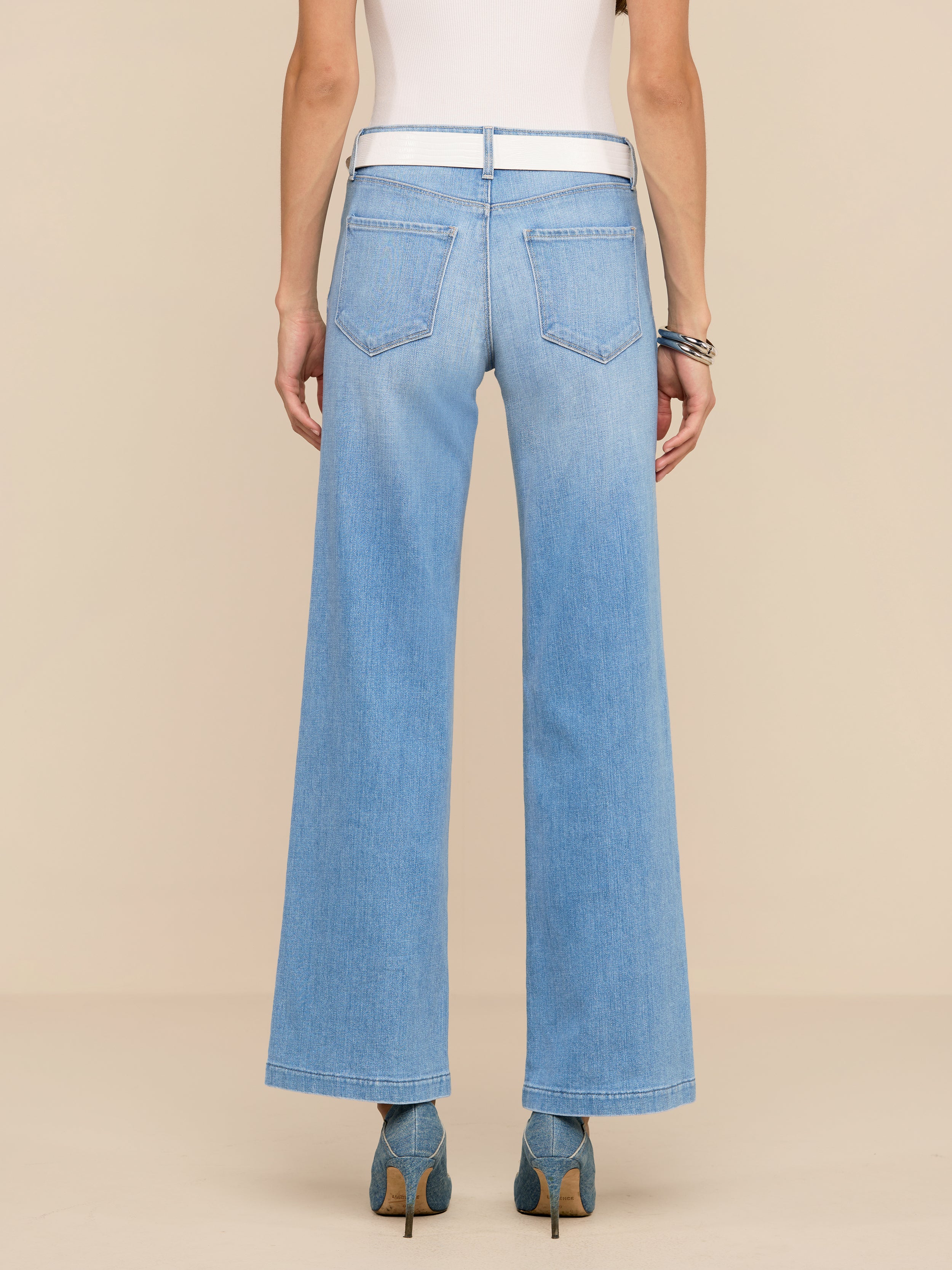 Une personne est représentée de dos sur un fond beige uni. Elle porte un jean Joanne Low-Rise Wide-Leg de couleur indigo clair, un haut blanc, des talons bleus et des bracelets.