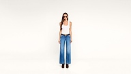 Woman wearing sunglasses, a white tank top, blue wide-leg jeans, and black boots stands against a plain white background, facing forward with a neutral expression.