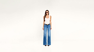 A woman wearing a white tank top, blue wide-leg jeans, a brown belt, and sunglasses stands against a plain background.
