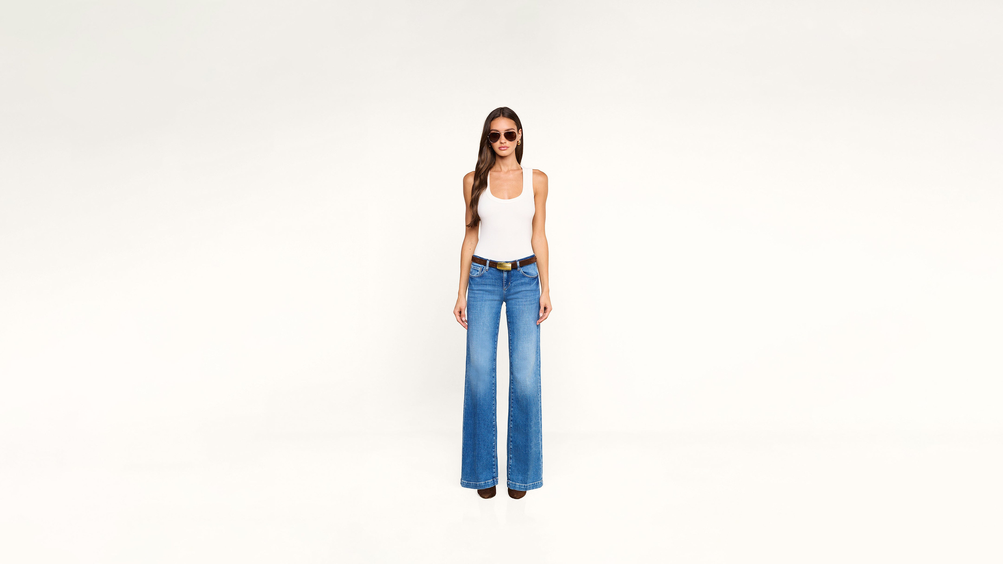 A woman wearing a white tank top, blue wide-leg jeans, a brown belt, and sunglasses stands against a plain background.