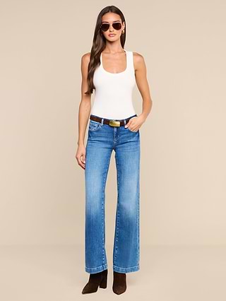 A woman wearing sunglasses, a white sleeveless top, the Joanne Low-Rise Wide-Leg Jean made from stretch denim, a black belt, and brown boots stands against a plain white background.
