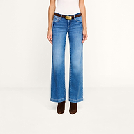 A person wearing the Joanne Low-Rise Wide-Leg Jean in blue stretch denim, styled with a black belt featuring a gold buckle, a white sleeveless top, and brown pointed-toe boots, stands against a plain white background.