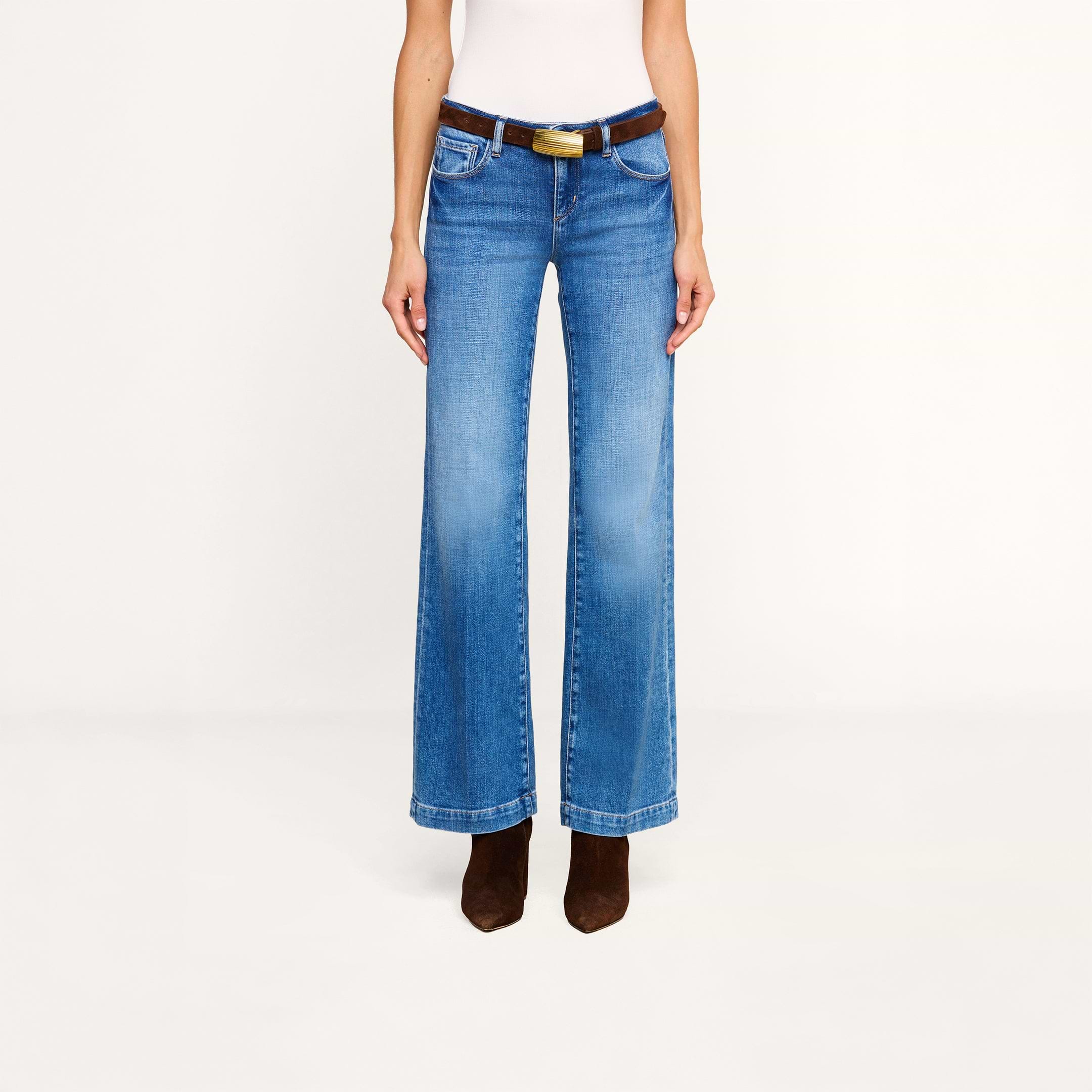 A person wearing the Joanne Low-Rise Wide-Leg Jean in blue stretch denim, styled with a black belt featuring a gold buckle, a white sleeveless top, and brown pointed-toe boots, stands against a plain white background.