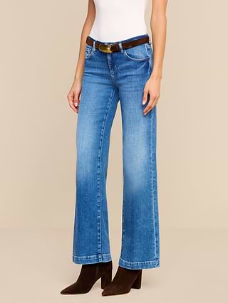 A person wearing a white top and the Joanne Low-Rise Wide-Leg Jean in blue stretch denim, styled with a brown belt and brown heeled boots, stands against a plain white background.