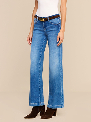 Model wears the Joanne Low-Rise Wide-Leg Jean in blue stretch denim, styled with a white sleeveless top, brown belt, and brown heeled boots. The model is standing against a plain beige background with their head not visible.