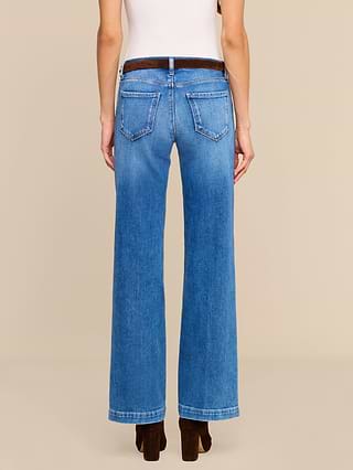A person stands facing away, wearing the Joanne Low-Rise Wide-Leg Jean in blue stretch denim, a white sleeveless top, and dark brown heeled shoes against a plain, light-colored background.