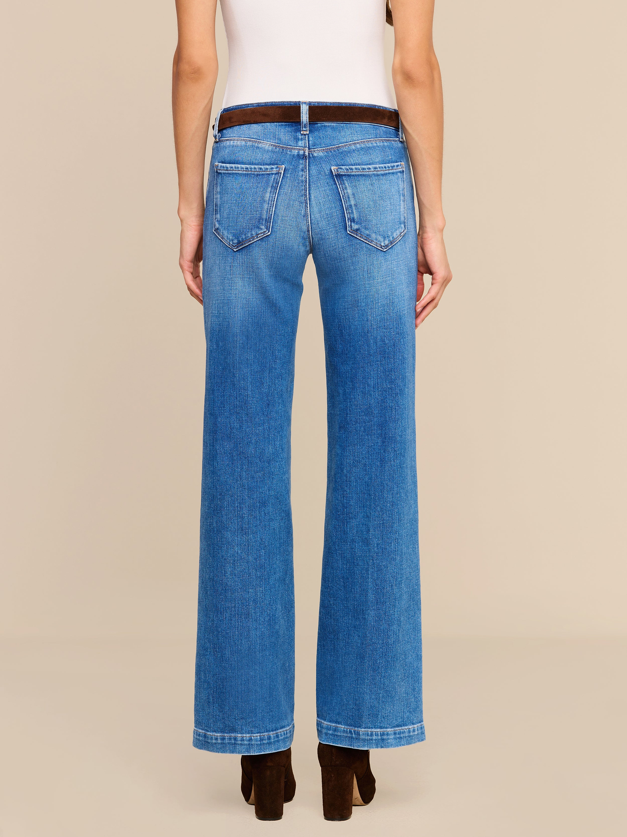 A person stands facing away, wearing the Joanne Low-Rise Wide-Leg Jean in blue stretch denim, a white sleeveless top, and dark brown heeled shoes against a plain, light-colored background.