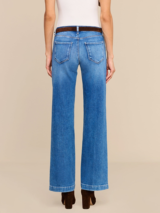 A person stands facing away, showing the back view, dressed in a white top, brown heeled shoes, and blue Joanne Low-Rise Wide-Leg Jeans in stretch denim.