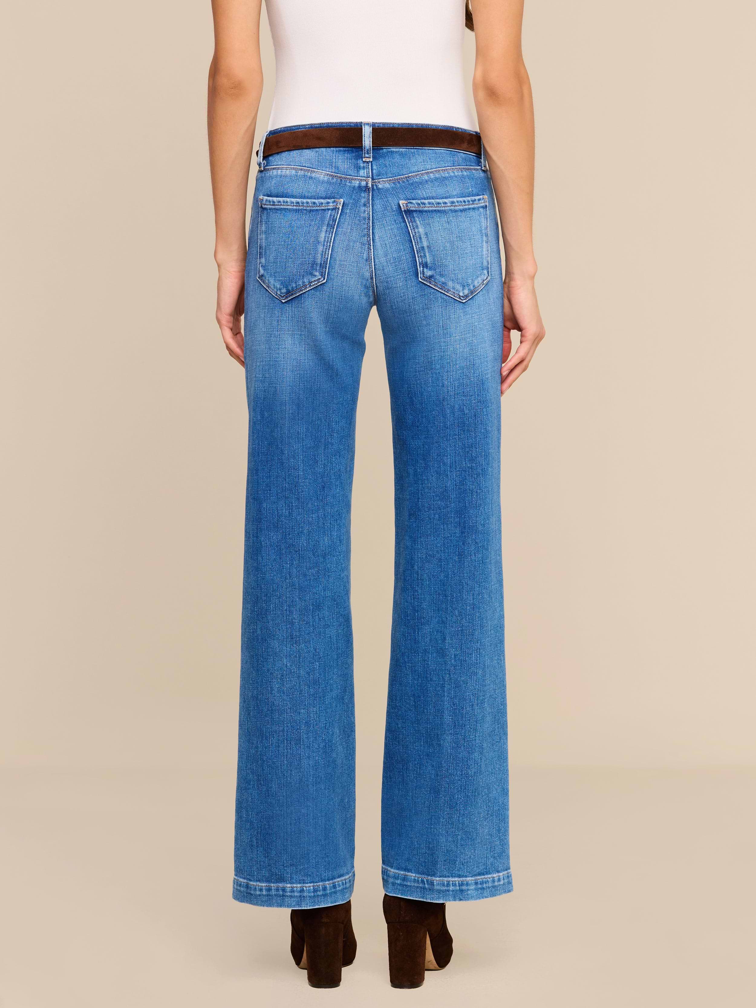 A person stands facing away, wearing the Joanne Low-Rise Wide-Leg Jean in blue stretch denim, a white sleeveless top, and dark brown heeled shoes against a plain, light-colored background.