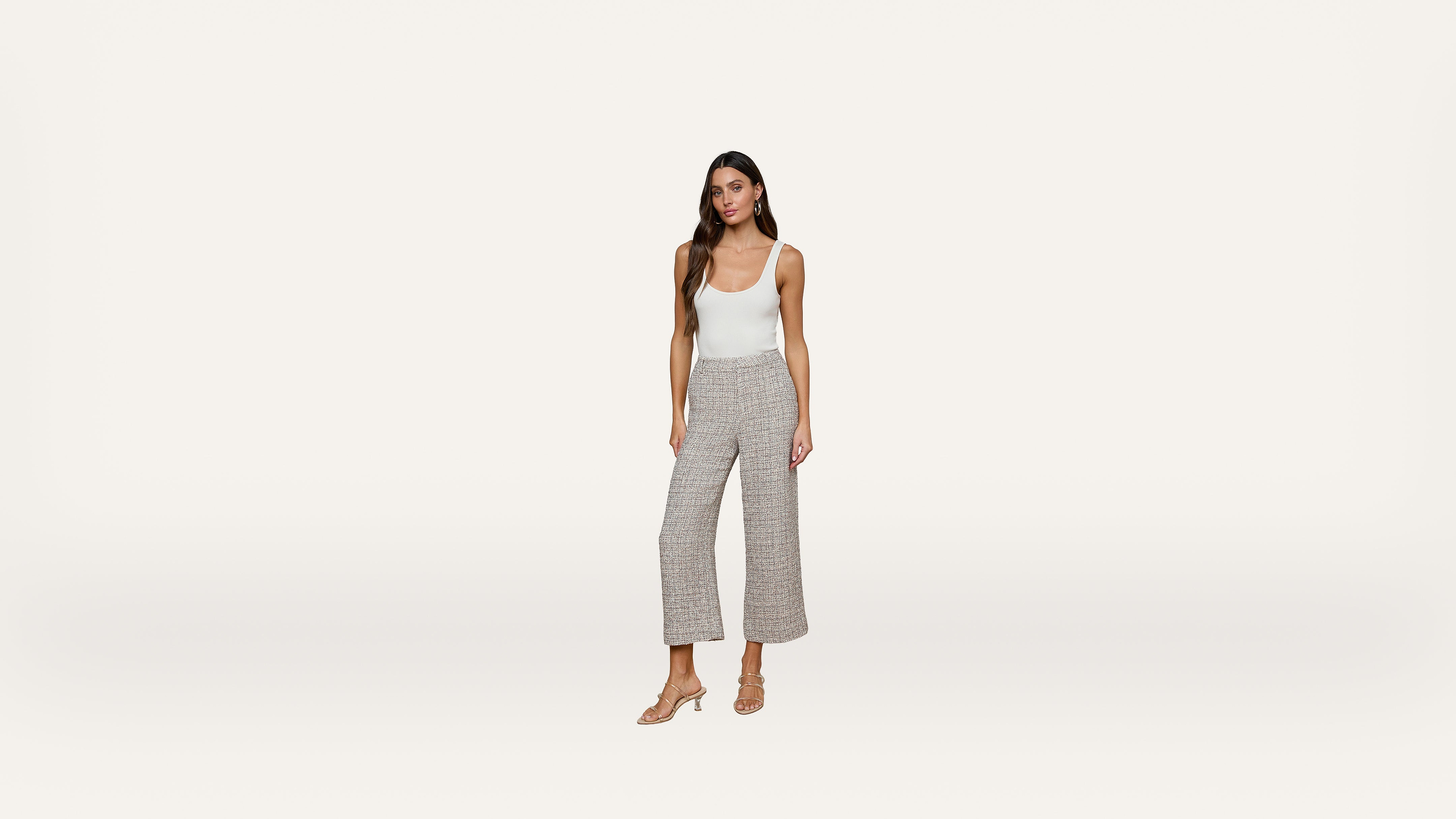 A woman stands against a plain white background wearing a white tank top, gray wide-leg pants, and beige heeled sandals.