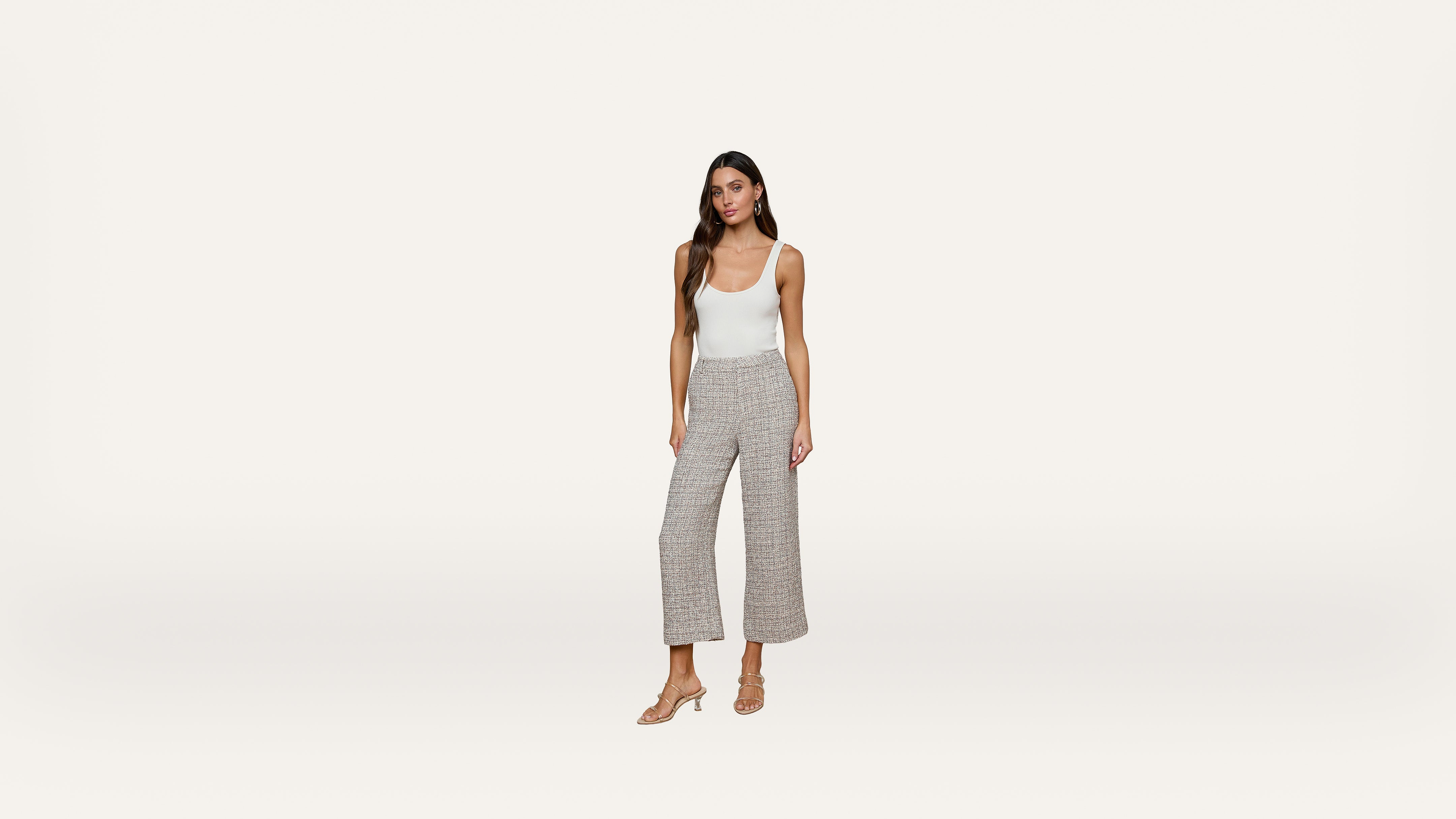 A woman stands against a plain white background wearing a white tank top, gray wide-leg pants, and beige heeled sandals.