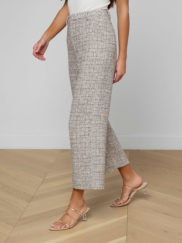 A woman wears the Lula Cropped Tweed Trouser, styled with clear heeled sandals and a partially visible white top, walking on a light wooden floor; her head is out of frame.