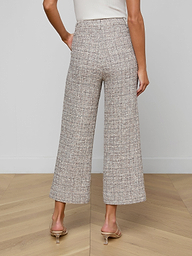 A woman stands on a wooden floor, facing away from the camera, wearing Lula Cropped Tweed Trousers, a white sleeveless top, and clear-heeled sandals.