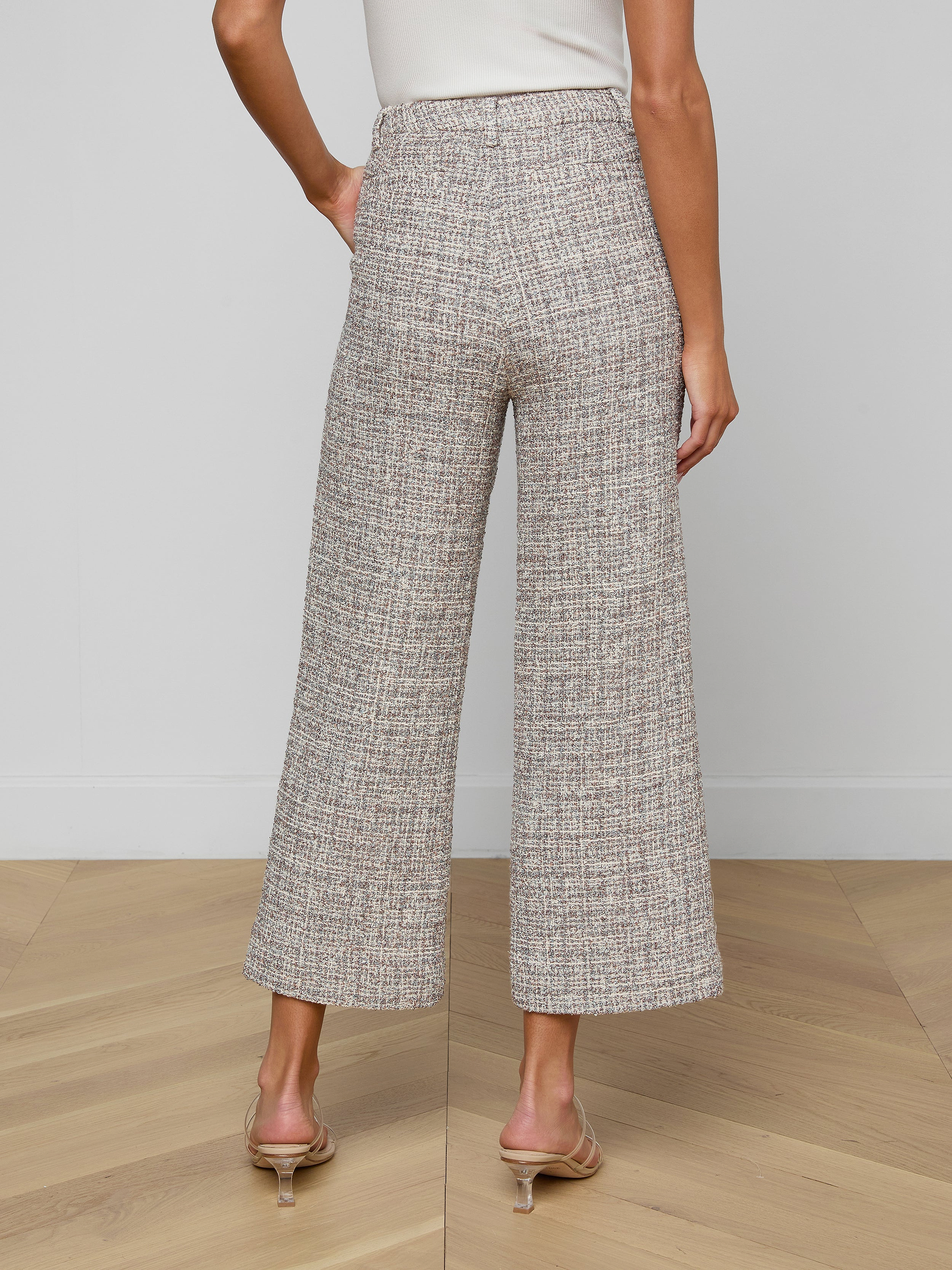A woman stands on a wooden floor, facing away from the camera, wearing Lula Cropped Tweed Trousers, a white sleeveless top, and clear-heeled sandals.