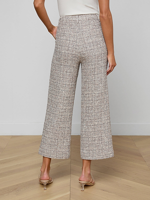 A woman stands on a wooden floor, facing away from the camera, wearing Lula Cropped Tweed Trousers, a white sleeveless top, and clear-heeled sandals.