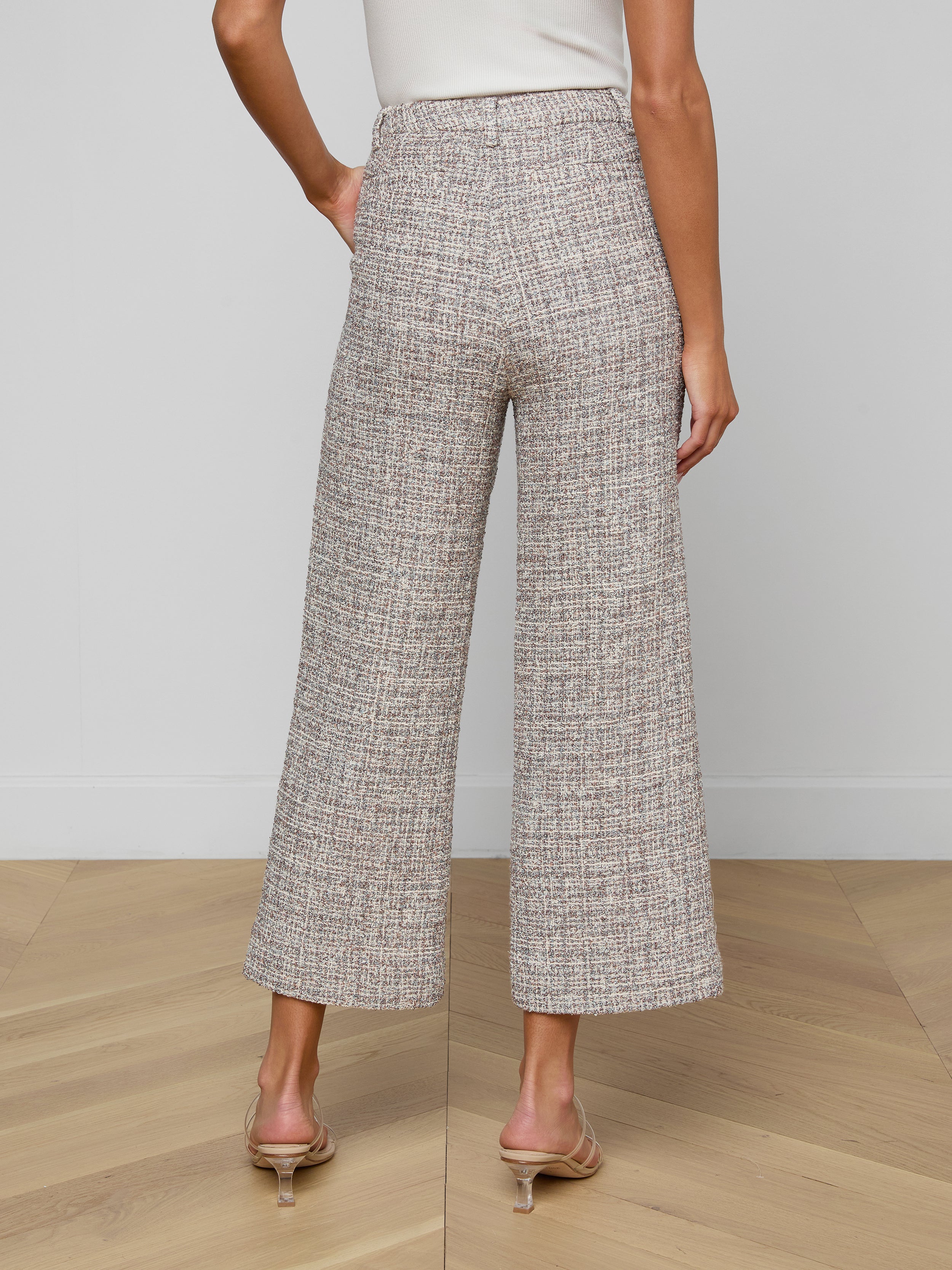 A woman stands on a wooden floor, facing away from the camera, wearing Lula Cropped Tweed Trousers, a white sleeveless top, and clear-heeled sandals.