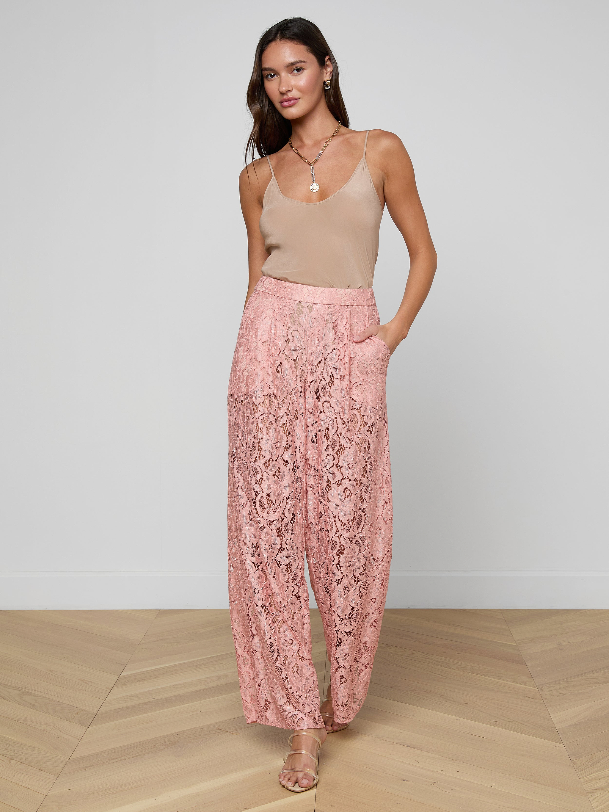A woman stands with one hand in her pocket against a plain white background, wearing a beige sleeveless top, heeled sandals, and the Tavia Lace Trouser—sheer stretch lace pink wide-leg pull-on trousers.