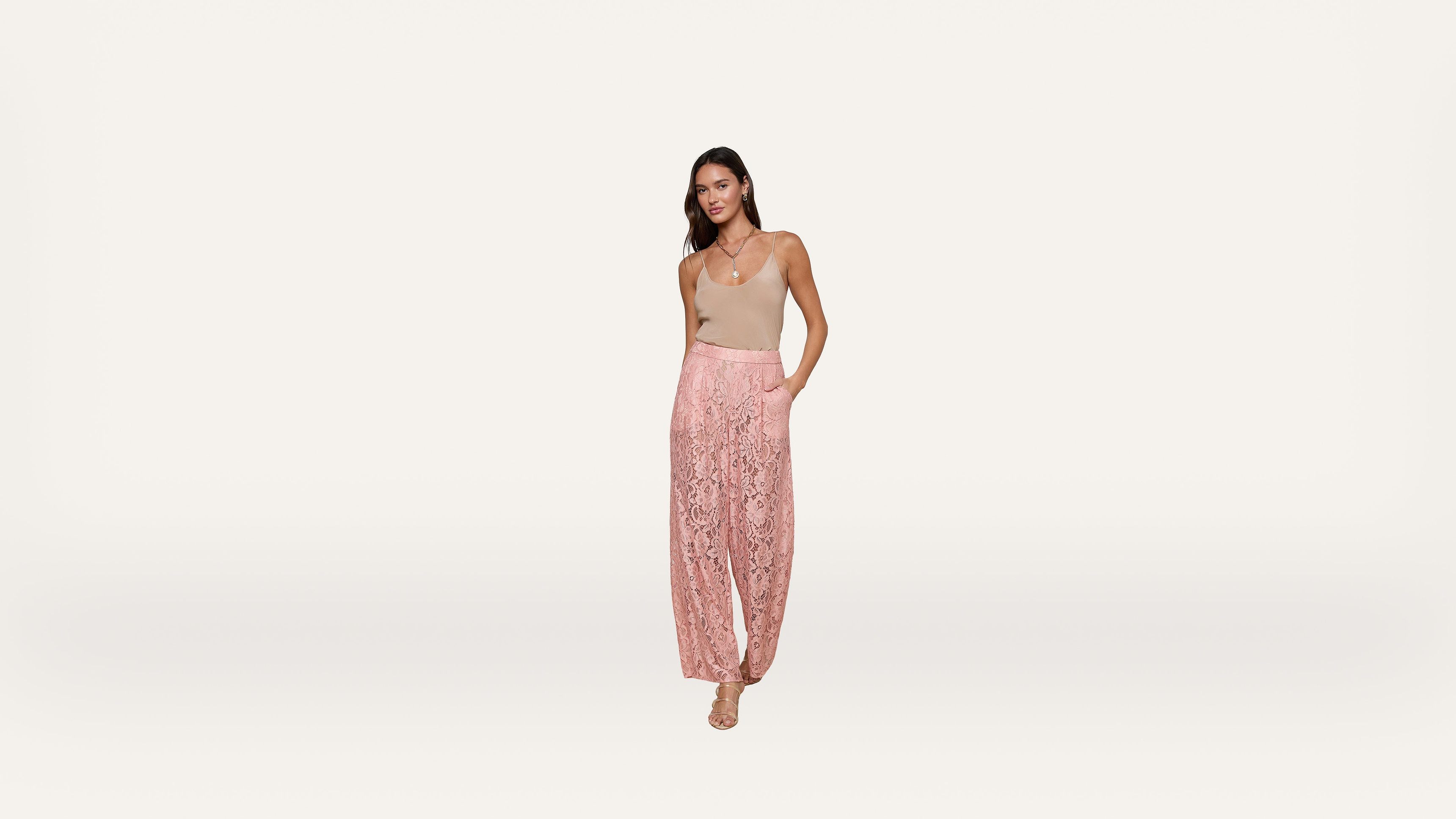 A woman stands against a white background, wearing a beige sleeveless top and high-waisted, wide-leg pink lace pants with pockets. She has long, dark hair and is wearing sandals.