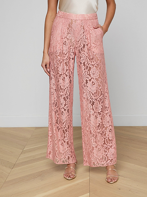 A woman stands on a wooden floor wearing the Tavia Lace Trouser—high-waisted, wide-leg pink sheer stretch lace pull-on pants—with a light satin top and clear-heeled sandals. Her hands are in her pockets; her face is not visible.