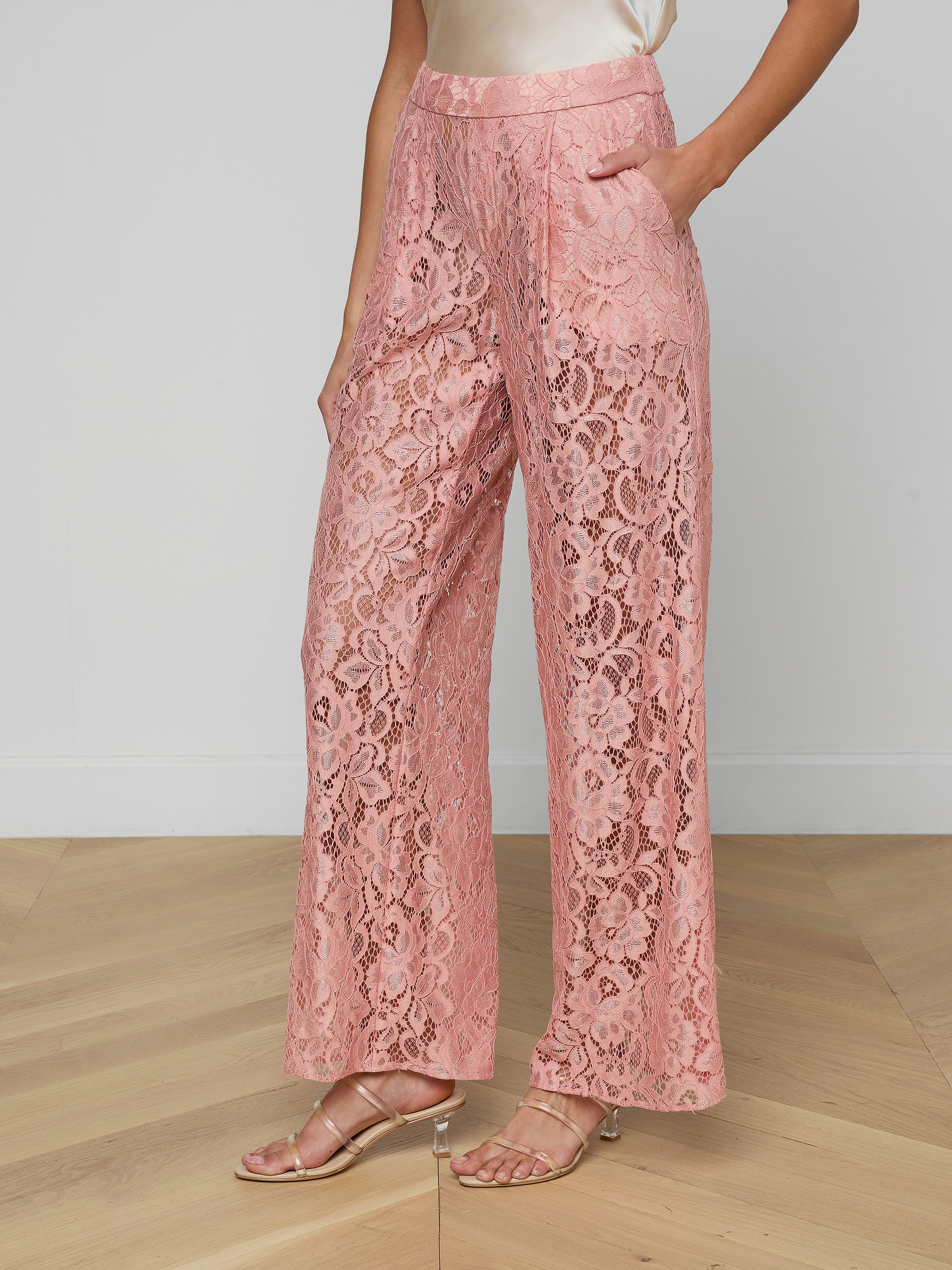A person wears the Tavia Lace Trouser—high-waisted, wide-leg, sheer stretch lace pull-on trousers in pink with front pockets—paired with heeled sandals and a sleeveless cream blouse, set against a plain, light-colored background.