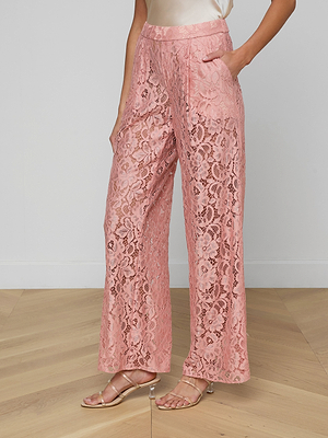 A woman stands on a wooden floor wearing blush pink Tavia Lace Trousers with pockets, paired with a cream satin sleeveless top and nude heeled sandals. Her left hand rests in her pocket.