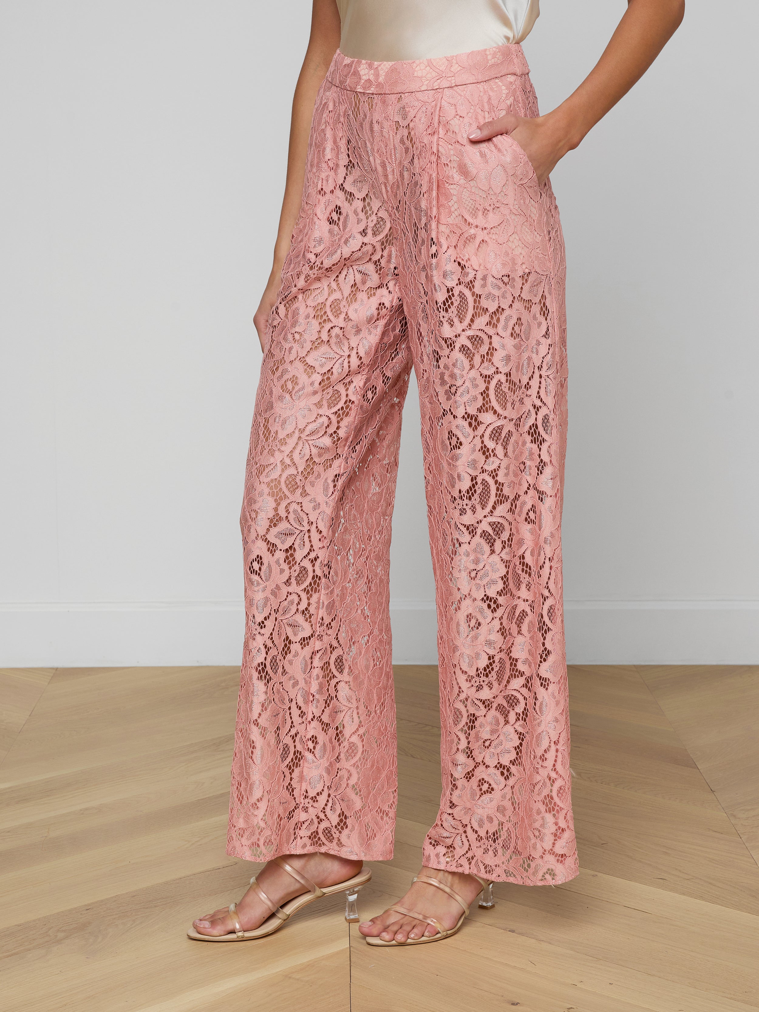 A person wears the Tavia Lace Trouser—high-waisted, wide-leg, sheer stretch lace pull-on trousers in pink with front pockets—paired with heeled sandals and a sleeveless cream blouse, set against a plain, light-colored background.