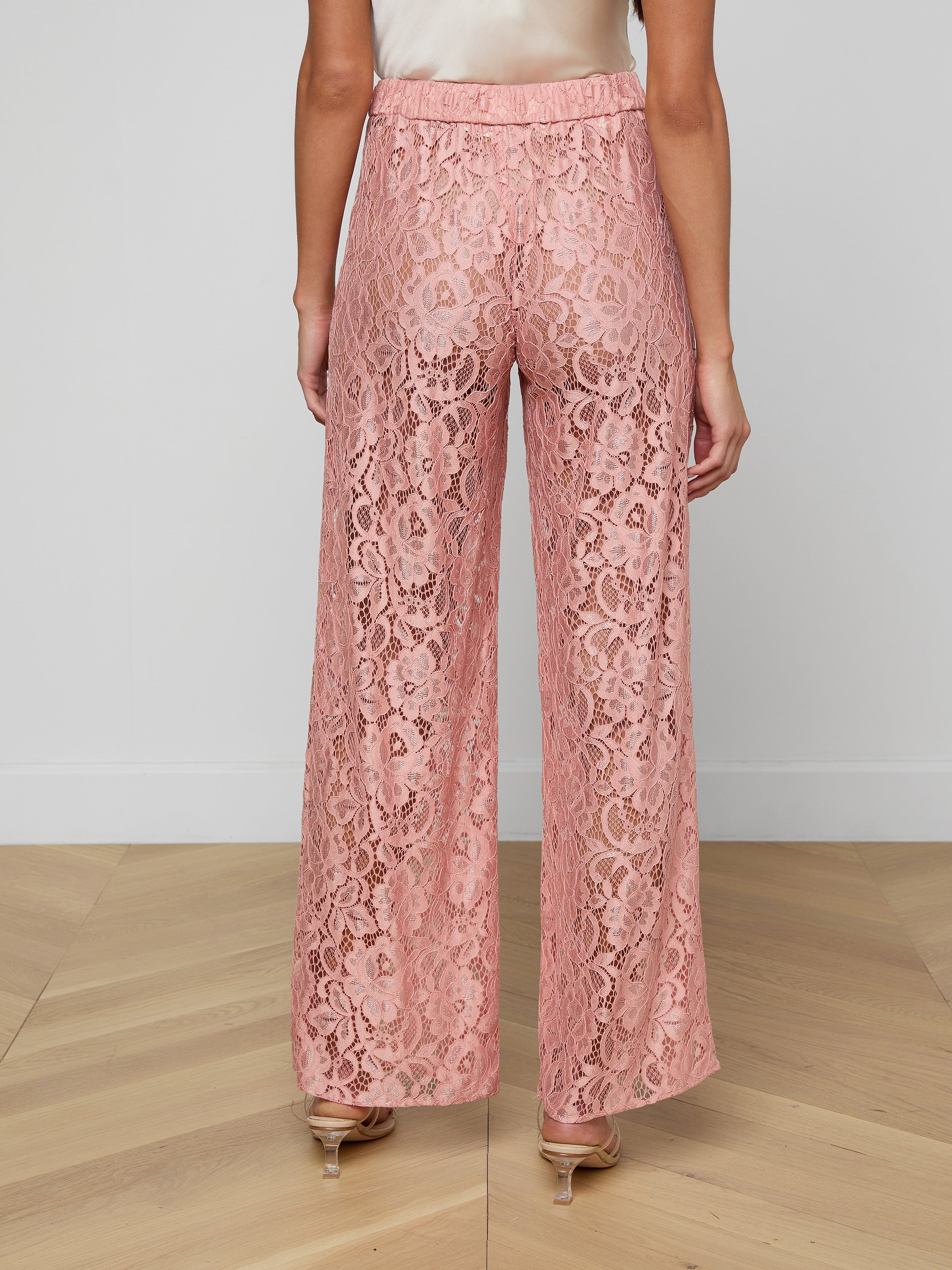 A woman is shown from the back against a plain white background wearing the Tavia Lace Trouser—high-waisted, wide-leg, pull-on trousers in sheer pink stretch lace—paired with beige heels.