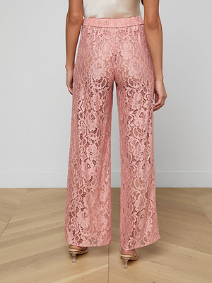 A woman stands indoors on a light wood floor, shown from the lower back down, wearing the blush pink Tavia Lace Trouser with sheer stretch lace, matching lining, and clear-heeled sandals.