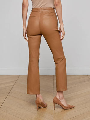 A person stands on a wooden floor, shown from the back, wearing the high-waisted tan Ketra Leather Bootcut Trouser and matching tan suede high-heeled shoes against a plain neutral background.