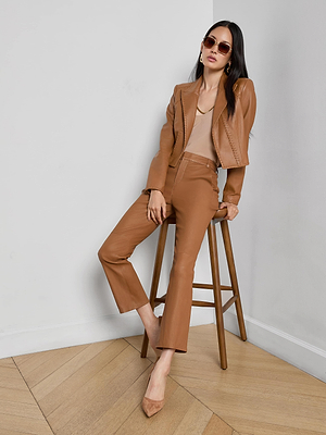A woman wearing sunglasses, a brown lambskin leather jacket, the Ketra Leather Bootcut Trouser, and tan heels sits on a wooden stool against a white wall and wooden floor, posing with one hand on her hip and the other resting on the stool.