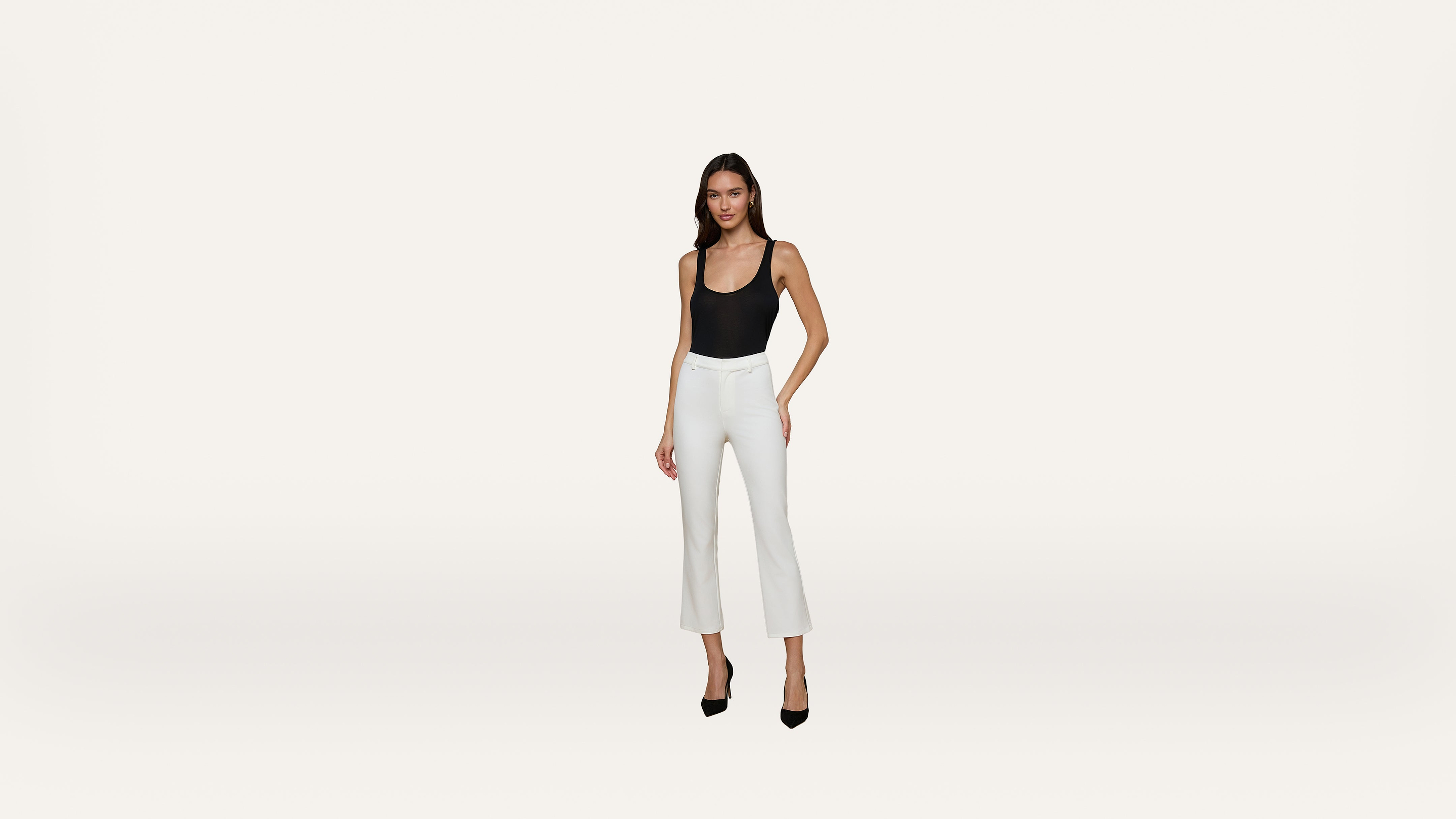 A woman wearing a black sleeveless top, white cropped pants, and black high heels stands against a plain white background.