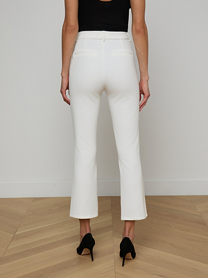 A person stands on a light wooden floor facing away from the camera, wearing white Ketra Bootcut Trousers in stretch suit fabric with black heels. The background is a plain, light-colored wall.