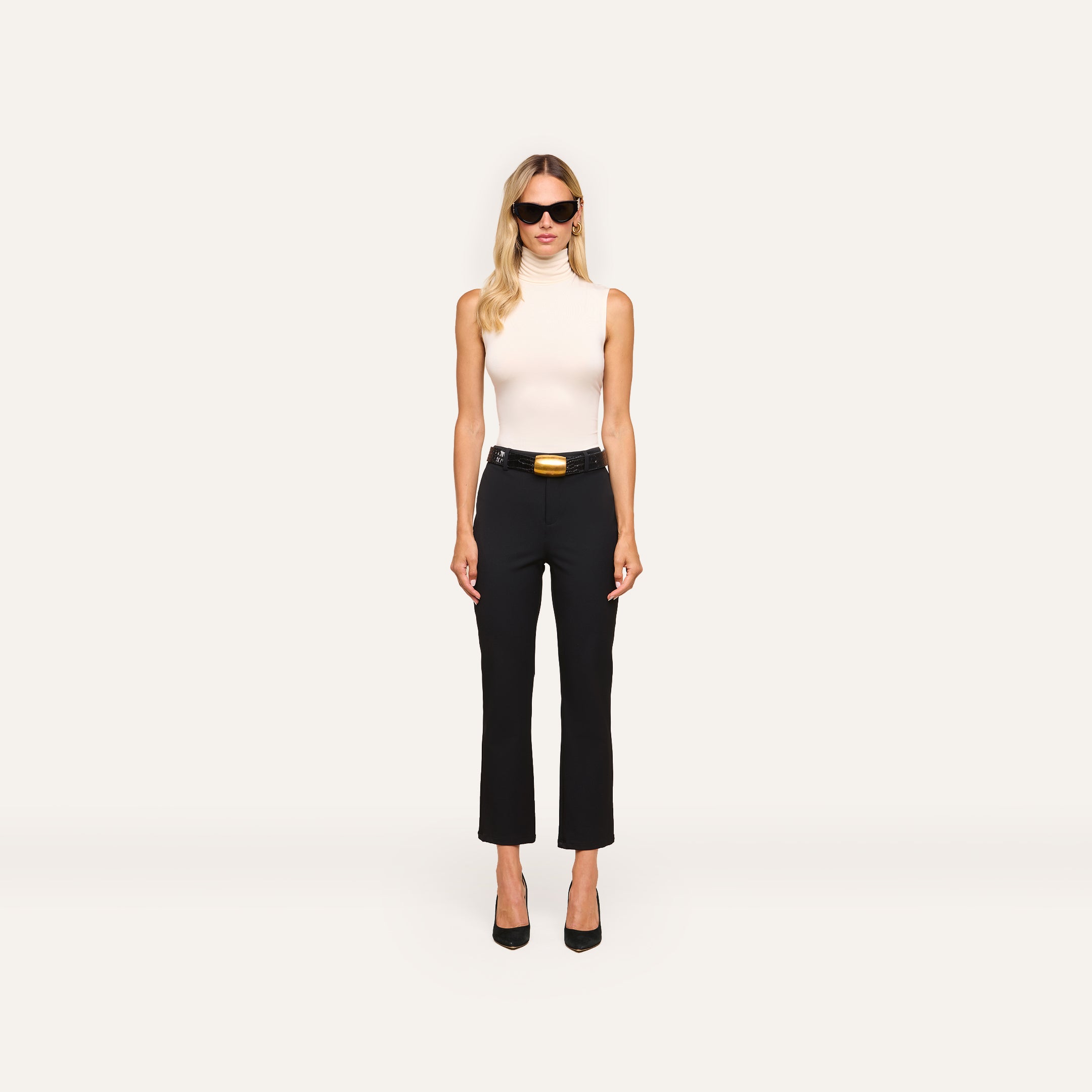 A woman with long blonde hair wears sunglasses, a white sleeveless turtleneck, and the Ketra Bootcut Trouser with a belt. She stands facing forward against a plain white background.