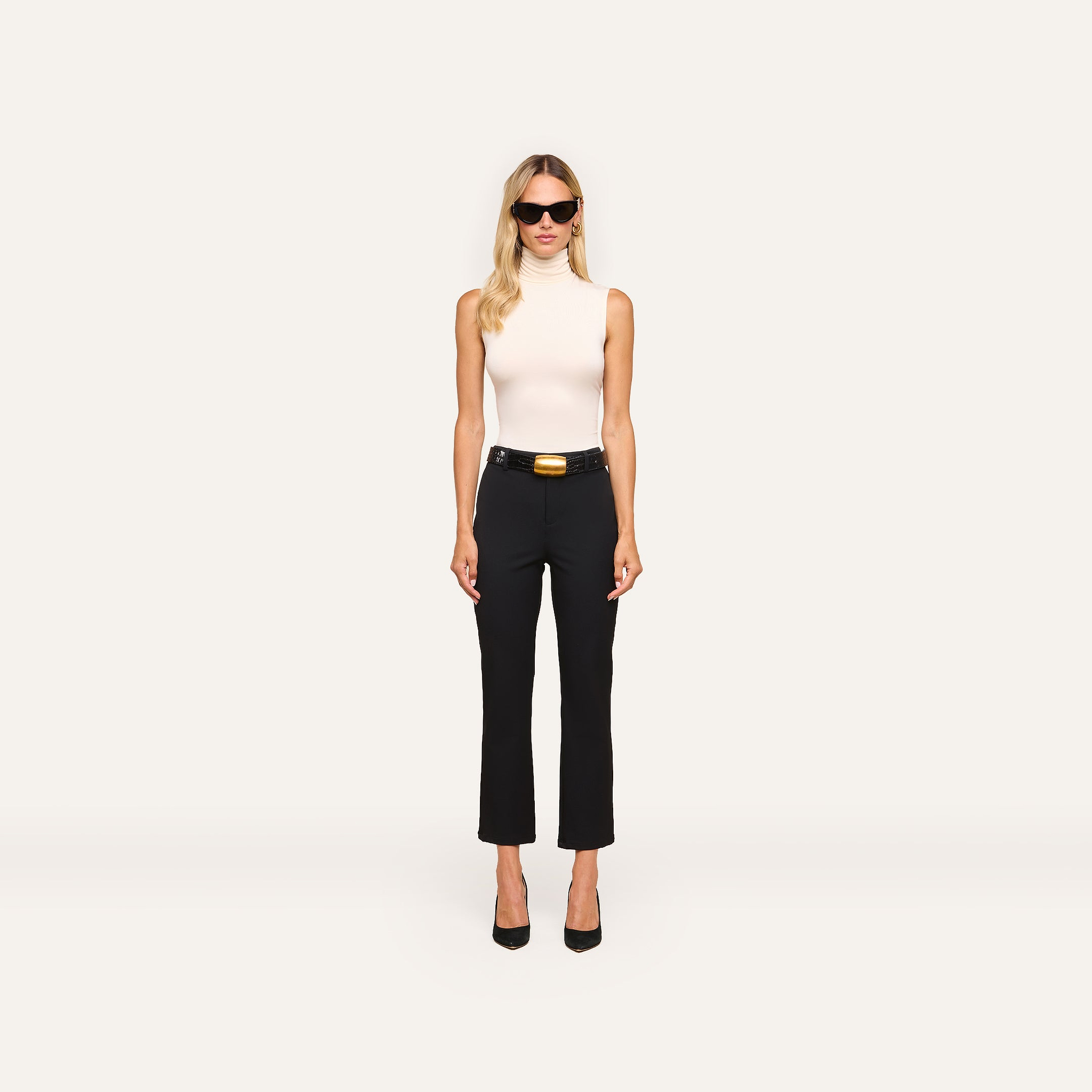 A woman with long blonde hair wears sunglasses, a white sleeveless turtleneck, and the Ketra Bootcut Trouser with a belt. She stands facing forward against a plain white background.