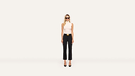 A woman with long blond hair wears sunglasses, a sleeveless white top, black pants, a black belt with a gold buckle, and black heels, posing stylishly against a plain white background.