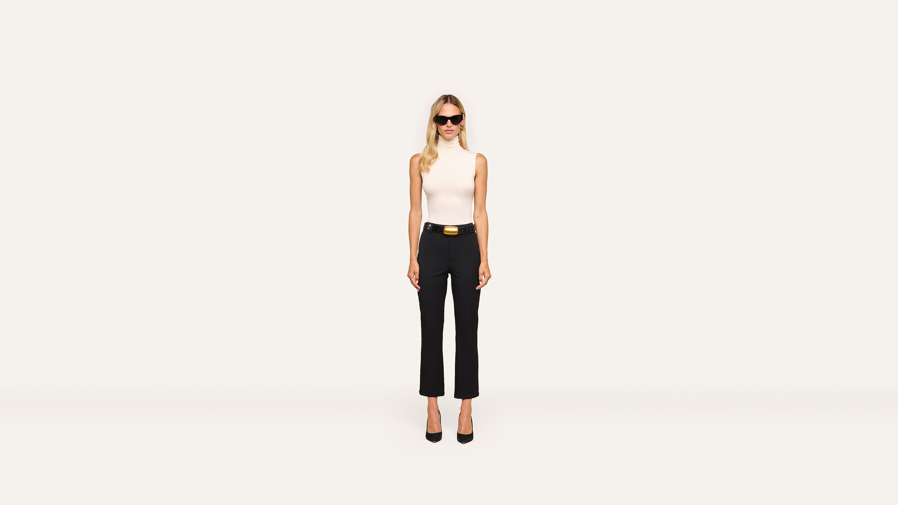 A woman with long blond hair wears sunglasses, a sleeveless white top, black pants, a black belt with a gold buckle, and black heels, posing stylishly against a plain white background.