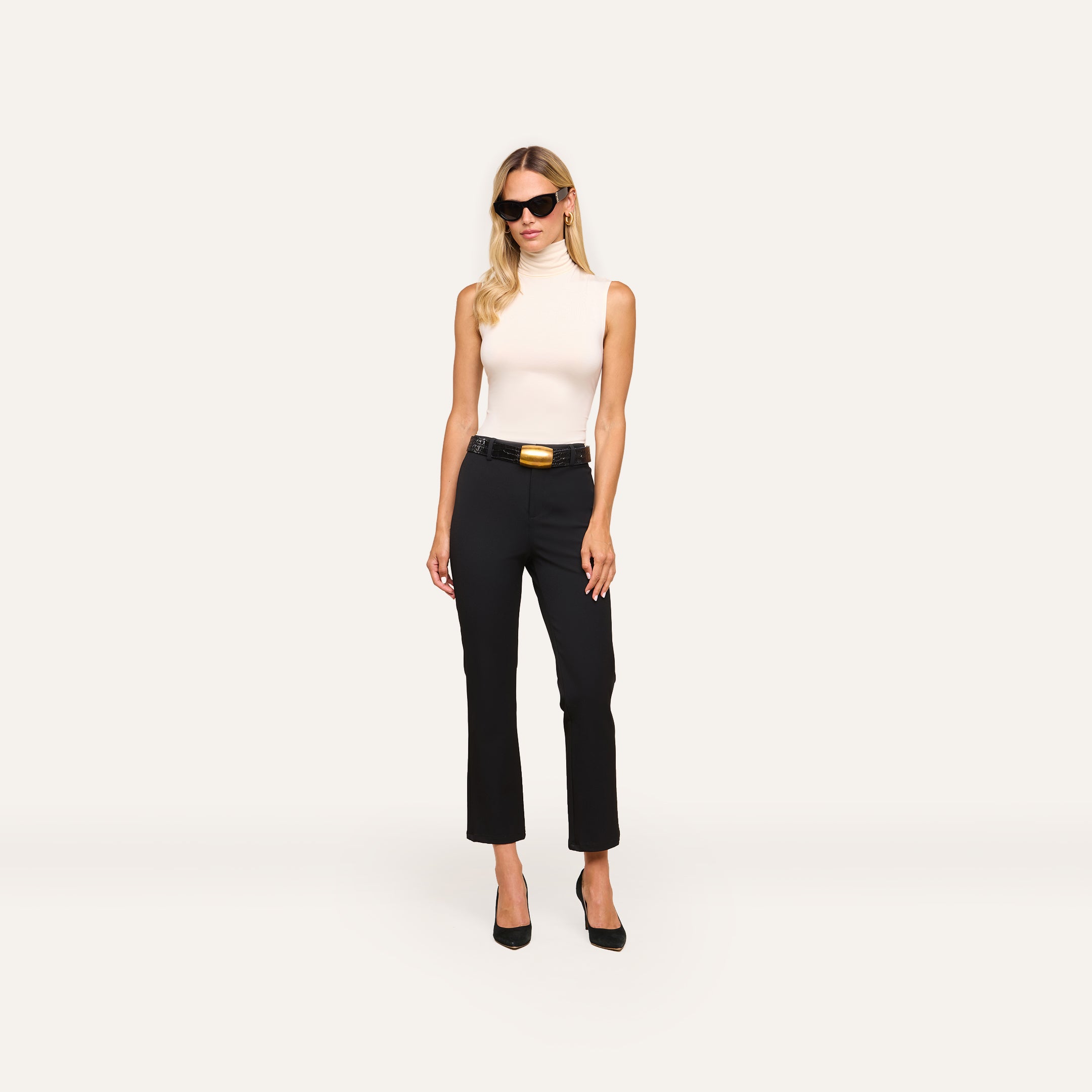 A woman with long blonde hair, sunglasses, a sleeveless white turtleneck, and the Ketra Bootcut Trouser with a wide belt and slant pockets stands against a plain light background.