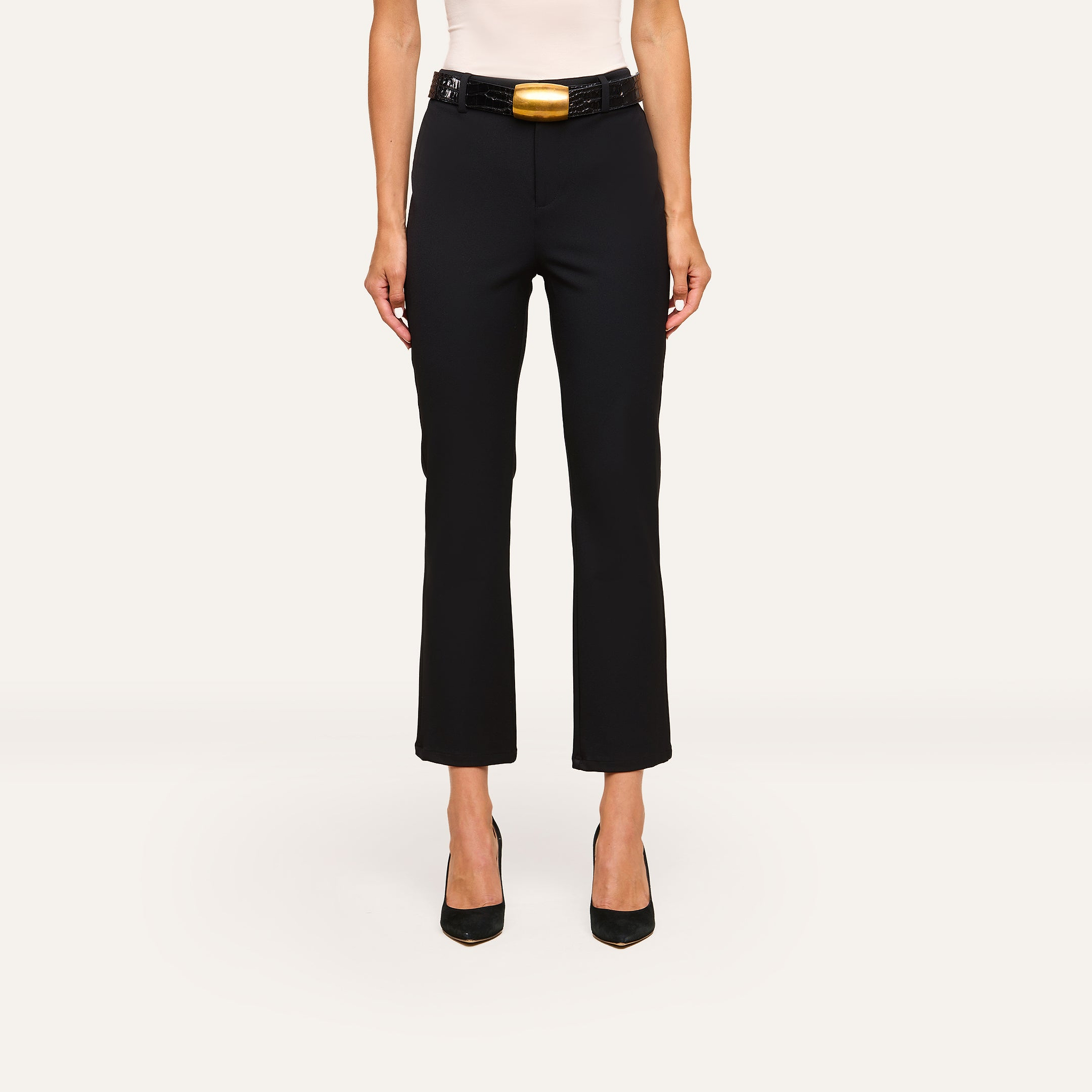 A woman, shown from the shoulders down, wears the Ketra Bootcut Trouser in black with slant pockets, a black belt with a large gold buckle, black heels, and a white top.