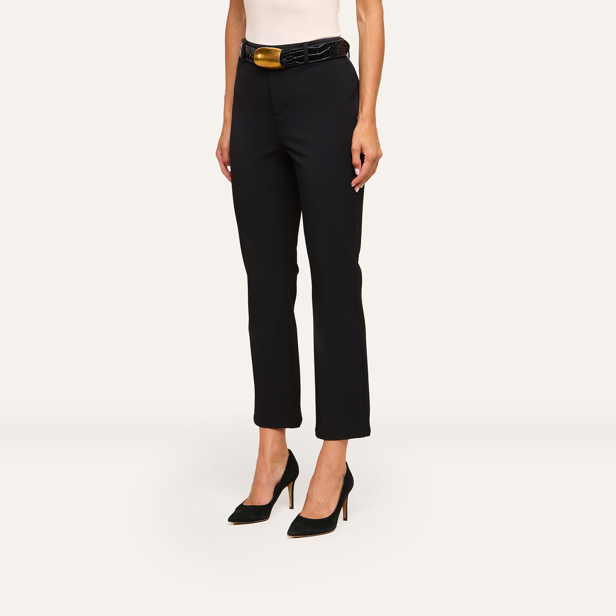 A woman wears Ketra Bootcut Trouser in black with slant pockets, paired with a wide black belt and black high heels. Only her lower torso and legs are visible against a plain off-white background.
