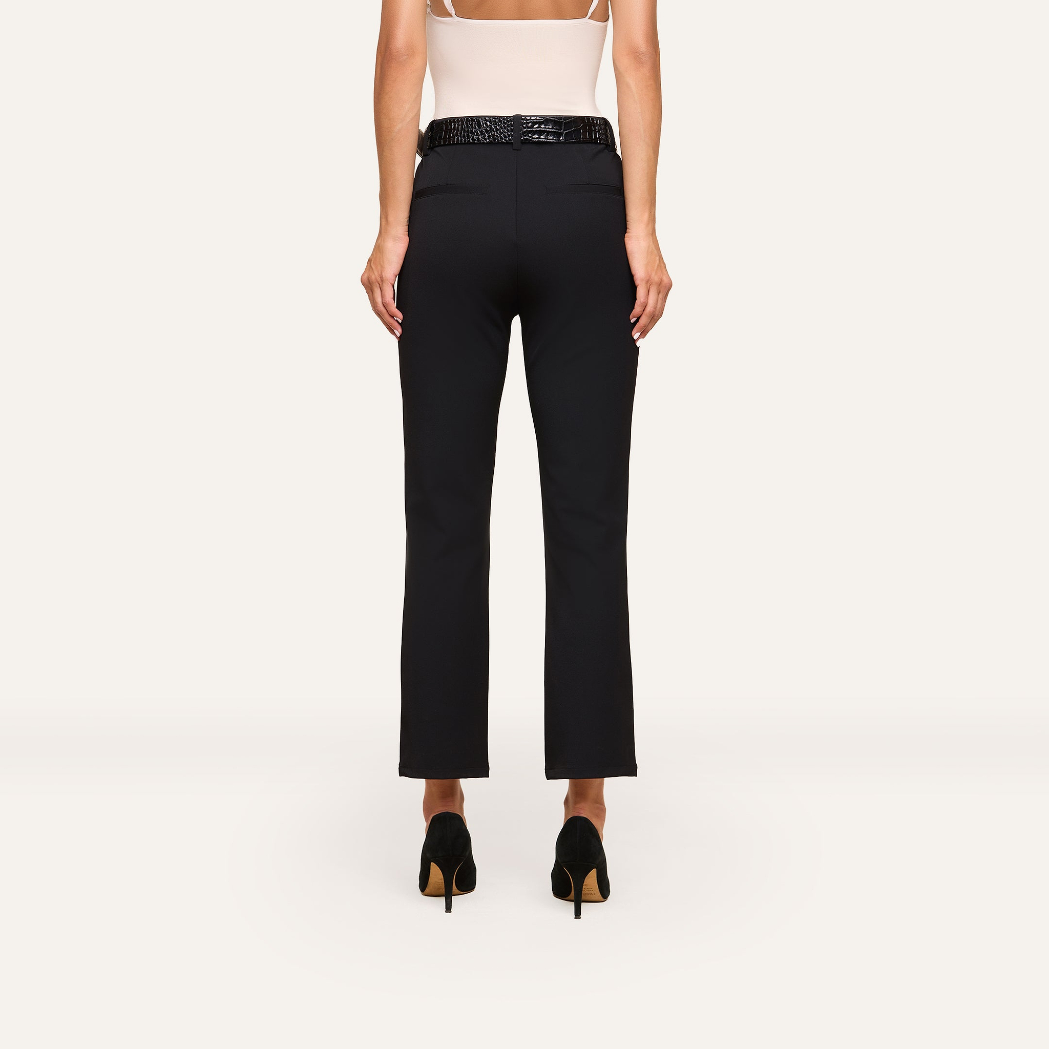 A woman stands facing away from the camera against a plain light background, wearing a white top and the Ketra Bootcut Trouser in black with slant pockets and a black belt.
