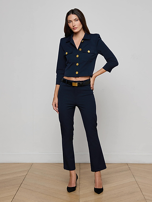 A woman with long brown hair stands indoors against a plain wall, wearing L'AGENCE's midnight navy Ketra Bootcut Trouser and a matching button-up jacket with gold buttons, paired with a black belt and black high heels.