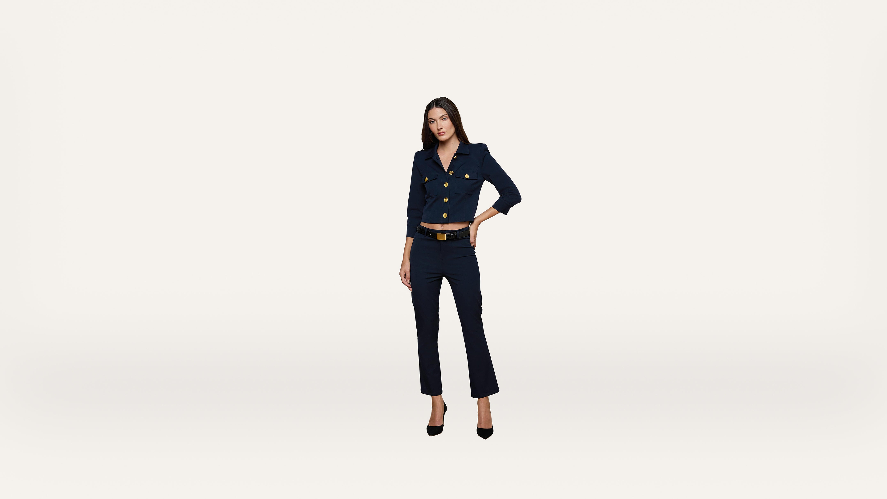 A woman in a navy blue suit with gold buttons stands confidently against a plain white background, her left hand on her hip and black heels on her feet.