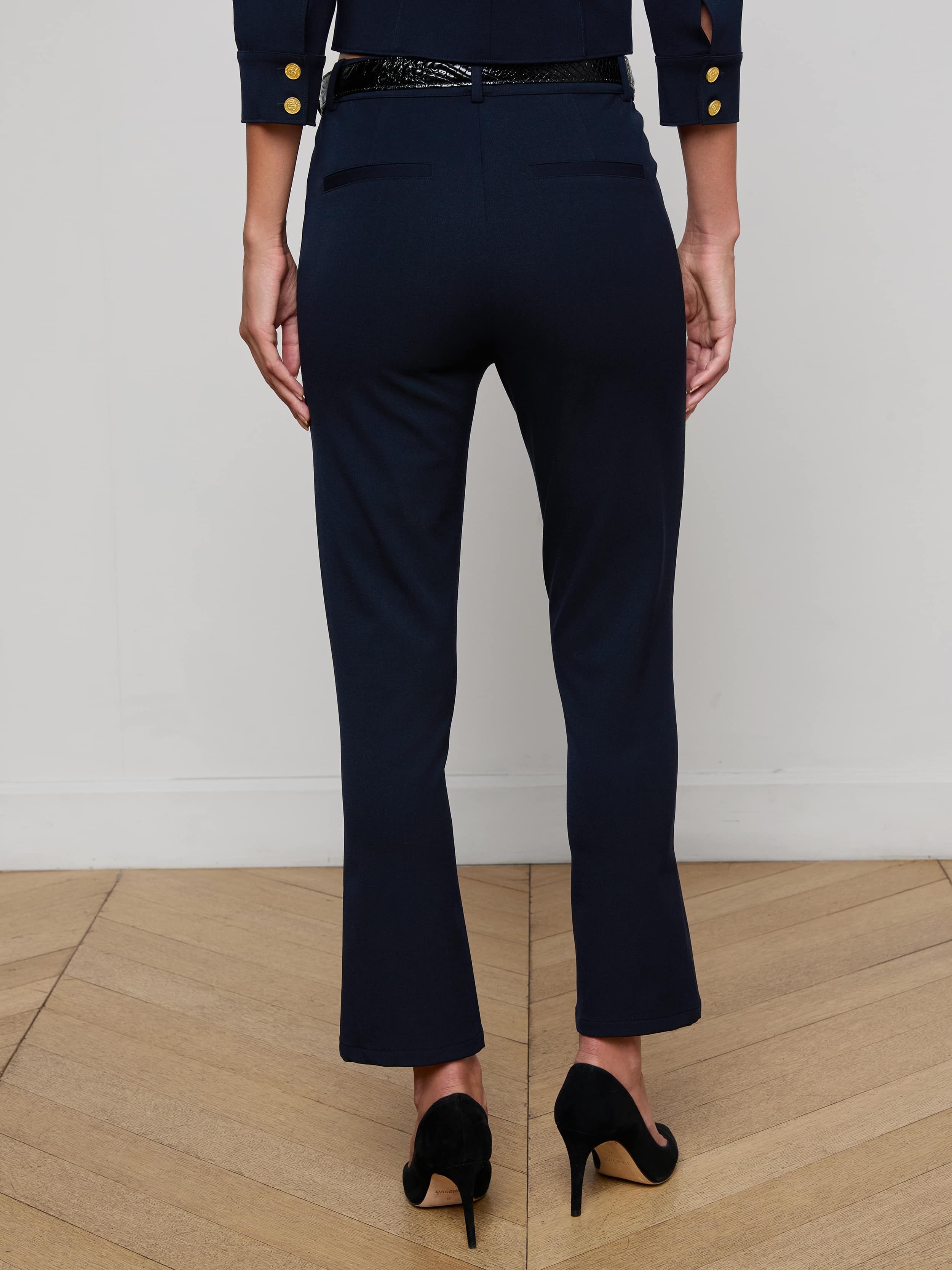 A person stands with their back to the camera, wearing the Ketra Bootcut Trouser in midnight navy and a matching blazer with gold buttons, both crafted from stretch suit fabric, paired with black high heels against a plain white background.