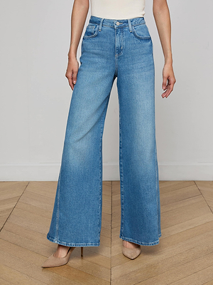 A person wears the L'AGENCE Beau Wide-Leg Jean in stretch blue denim, a sleeveless white top, and beige pointed-toe heels while standing on a wooden floor against a white wall.