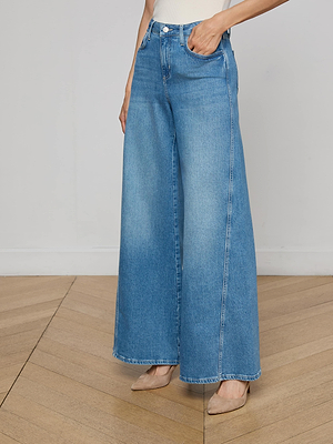 A person wearing L'AGENCE Beau Wide-Leg Jeans in blue with beige pointed-toe heels stands on a light wood floor against a plain white wall; their upper body is mostly out of frame.