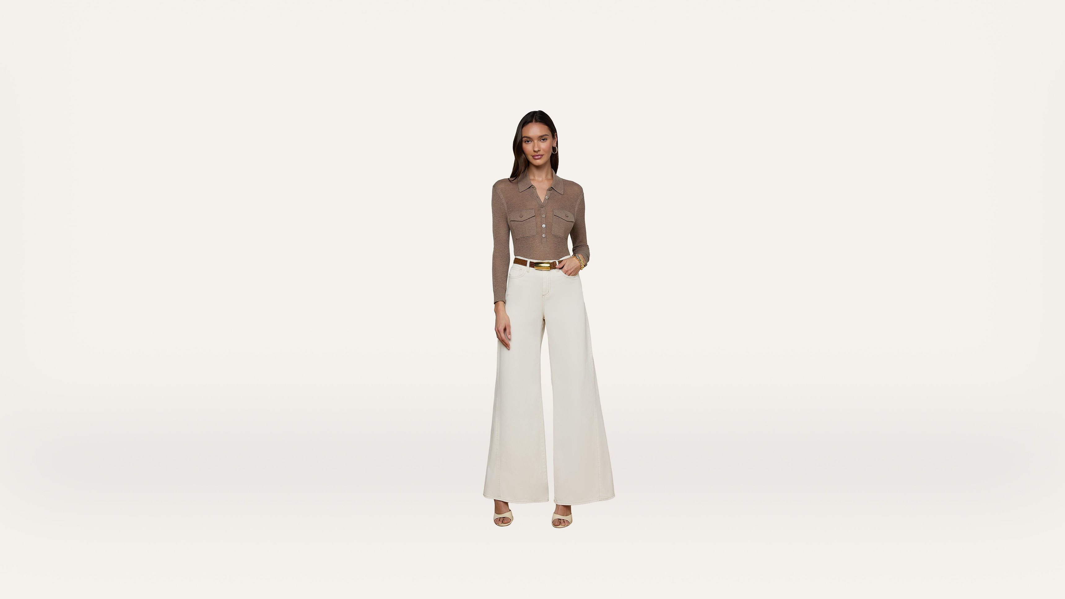 A woman stands against a plain white background, wearing a brown long-sleeve top, wide-leg white pants, a tan belt, and open-toe heels.