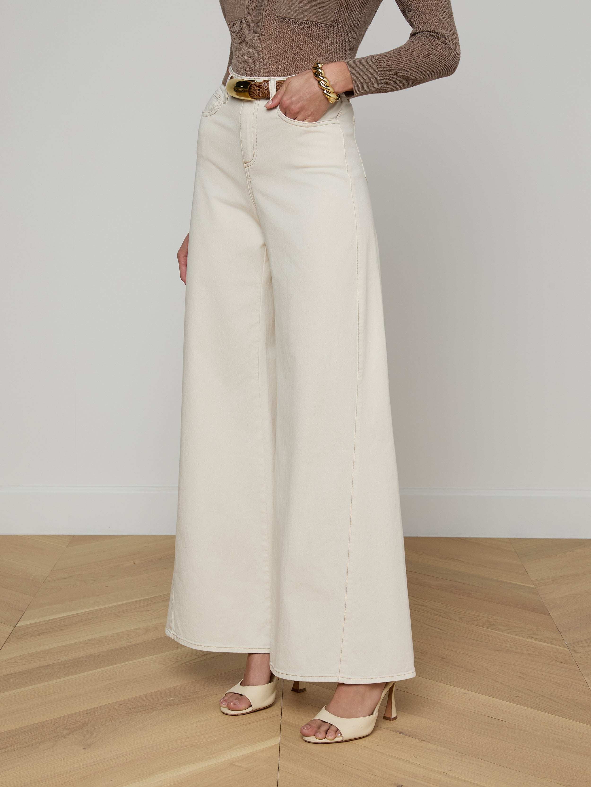 A person stands against a plain light background wearing the Beau Wide-Leg Jean—a high-rise, wide-leg style in white stretch cotton twill. Only the lower half of their body is visible, styled with beige heeled sandals, a belt, and a bracelet.