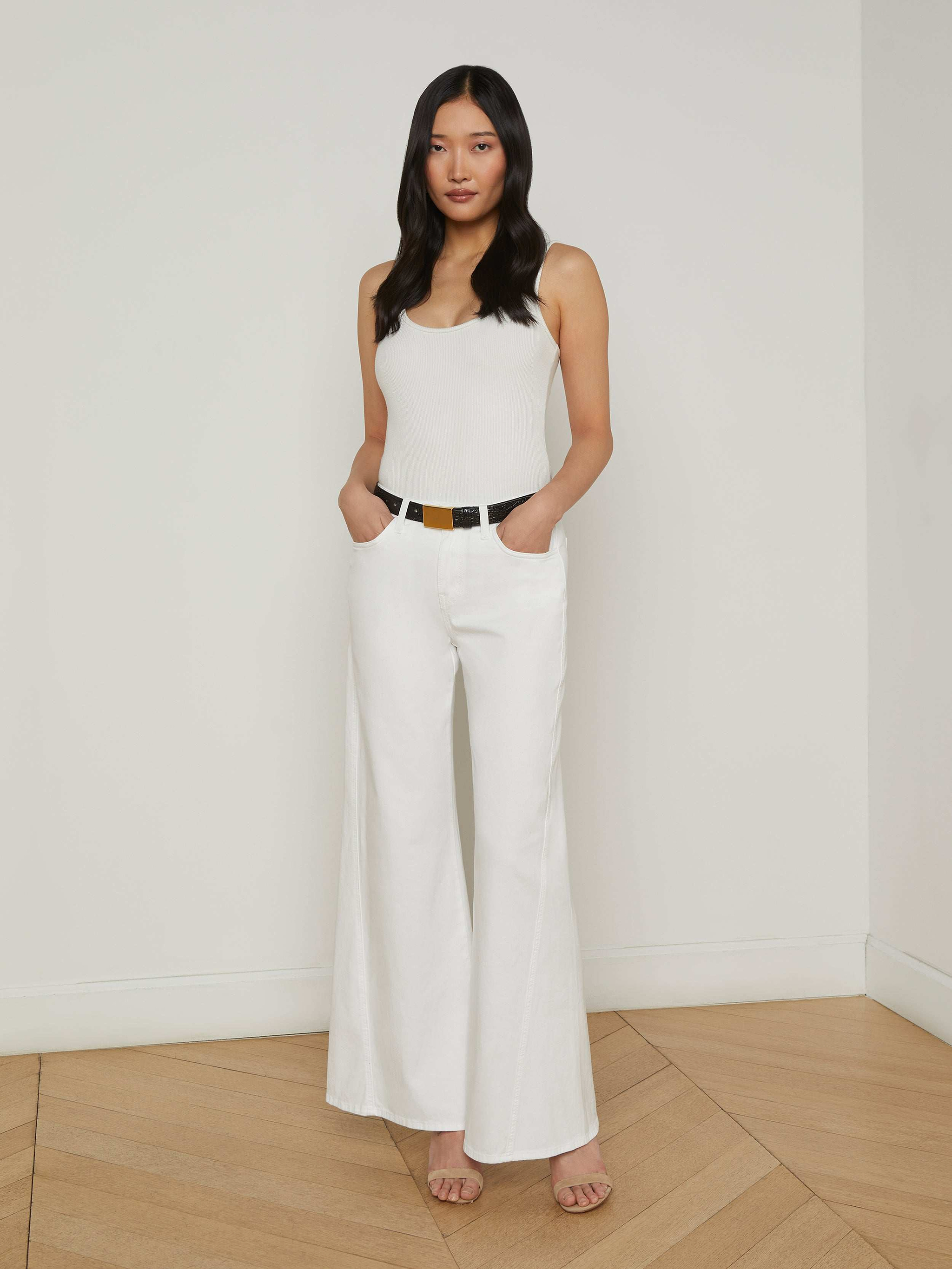 A woman stands against a plain white background wearing a white tank top, the Beau Wide-Leg Jean in high rise stretch denim, a black belt, and beige heels, with her hands in her pockets.