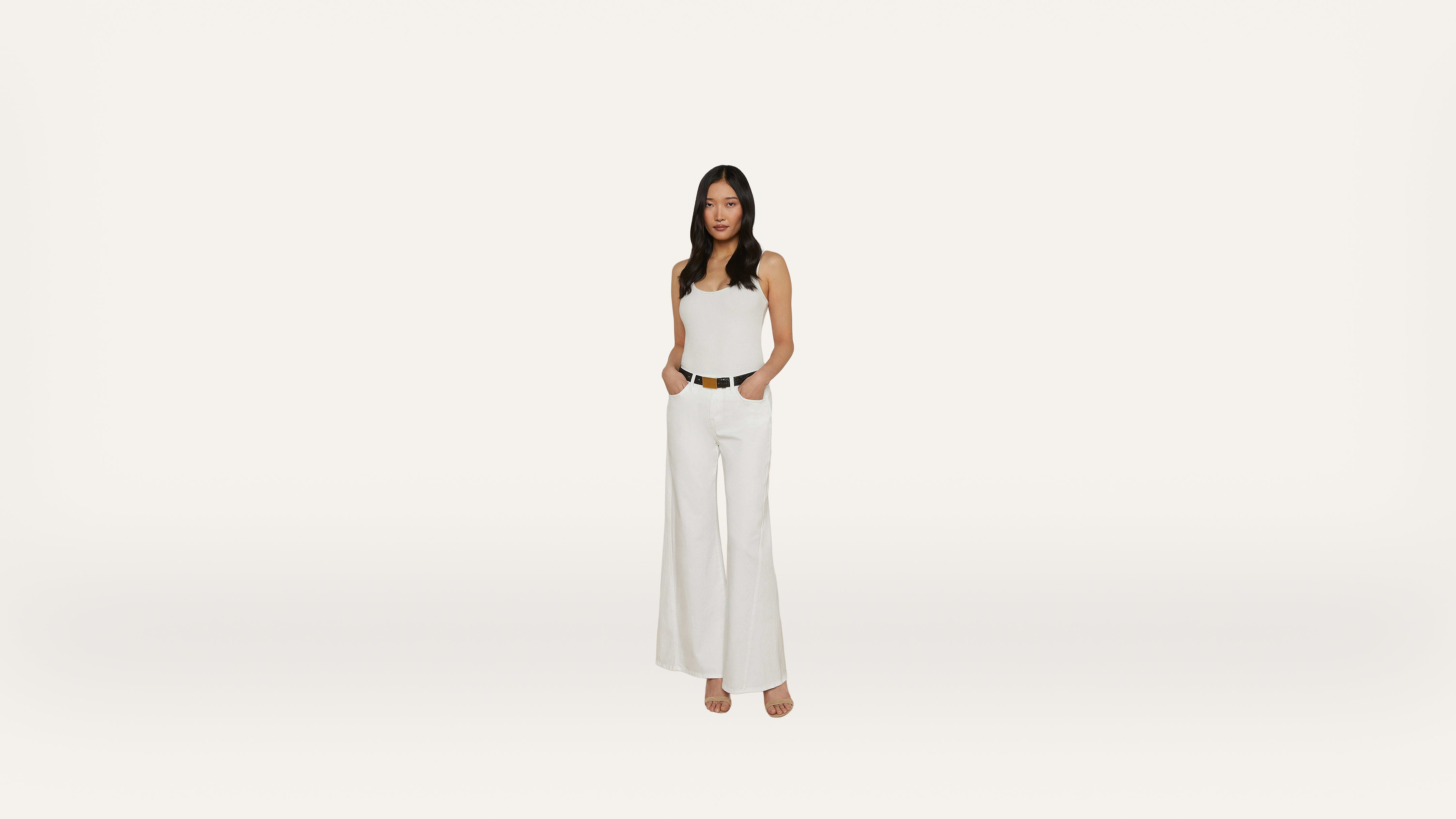 Woman wearing a white sleeveless top and wide-leg white pants, standing with hands in pockets against a plain white background.